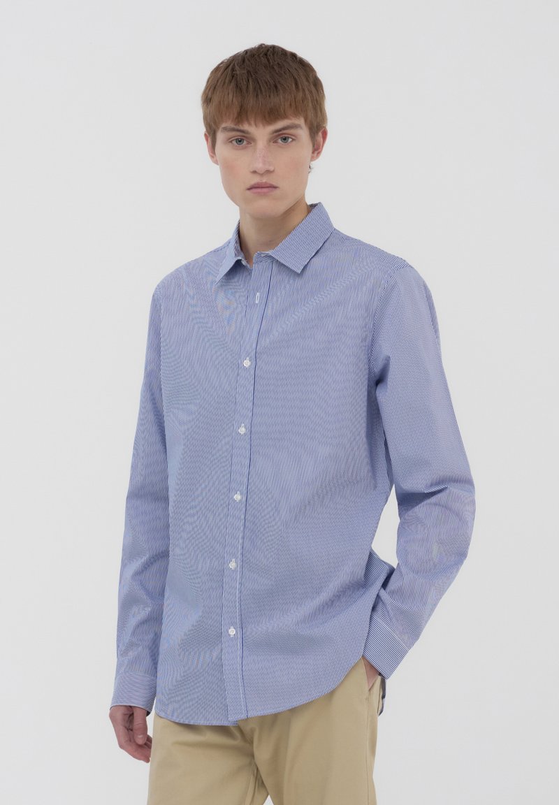 Camicia azzurro chiaro con sottili strisce bianche, colletto classico e maniche lunghe, realizzata in un tessuto liscio. Indossata con pantaloni beige.