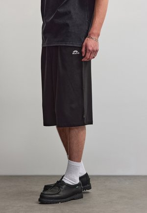 Mann trägt schwarze knielange Shorts mit weißem Logo, schwarze Schuhe, weiße Socken, schwarzes T-Shirt, steht auf grauem Boden vor grauer Wand.