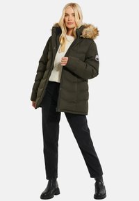 Khaki pufferjack met een nep-bont capuchon, ritssluiting en gewatteerde textuur. Ggedragen over een crèmekleurige trui en zwarte broek met zwarte laarzen.