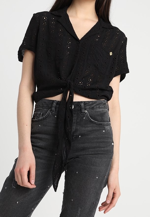 Maglietta nera a maniche corte con nodo frontale, motivo ad occhio di pernice e una tasca sul petto. Abbinata a jeans scuri decorati con borchie trasparenti.