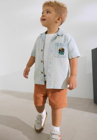 Camisa de mezclilla de manga corta en azul claro con botones de madera y un bolsillo que presenta un diseño bordado colorido; combinada con pantalones cortos naranjas y zapatillas.