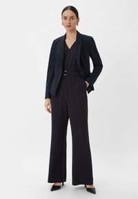 Marineblauer Blazer über einem ärmellosen Jumpsuit mit tiefem V-Ausschnitt. Der Jumpsuit hat eine taillierte Taille mit einem Gürtel und weiten Hosenbeinen. Schwarze Absätze.