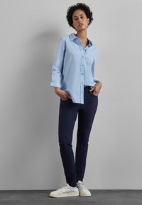 Světle modrá košile s rukávy na ohrnutí, spárovaná s námořnickými slim-fit kalhotami a bílými teniskami na šedém pozadí.