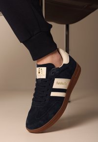 Granatowe zamszowe trampki z białymi pasiakami, kremowymi akcentami i brązową gumową podeszwą. Mają logo na języku oraz okrągły kształt noska.