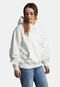 Felpa bianca oversize realizzata in tessuto morbido, presenta una tasca a marsupio, polsini a coste e un piccolo logo sul petto. Abbinata a jeans blu.