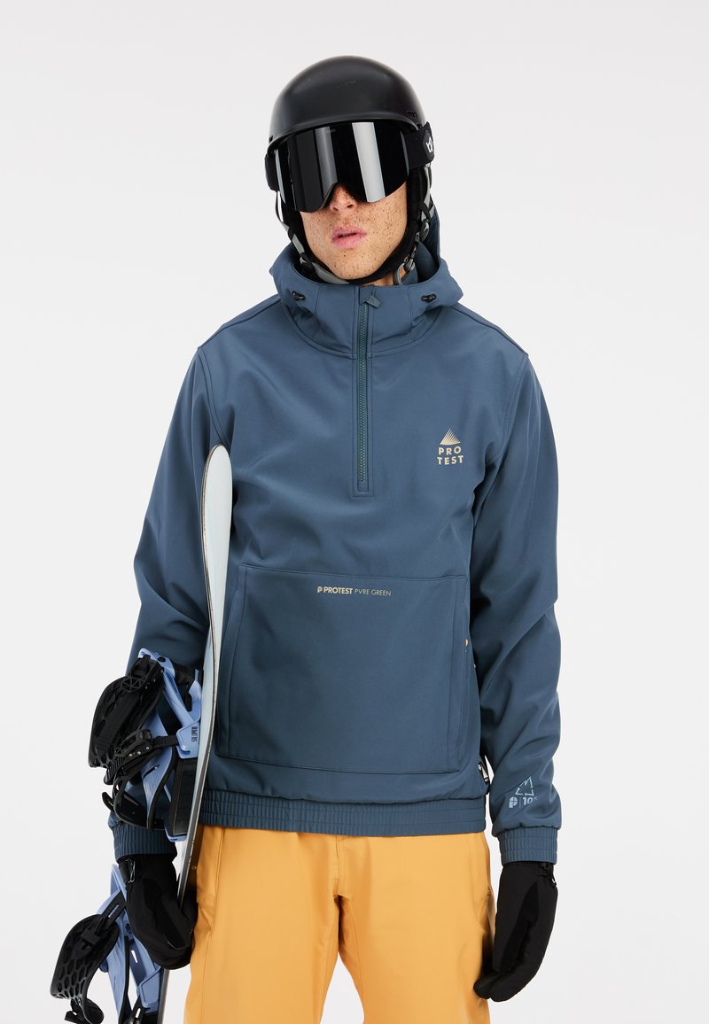 Dark blue snowboard jacket with gold logo accents, zippered pocket, high collar, and adjustable cuffs. Model holds a snowboard.