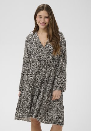 Mlada ženska, ki se smeji, oblečena v črno-belo obleko z živalskim vzorcem zebre z dolgimi rokavi in do kolen ter sproščenim krojem, na enobarvnem ozadju.