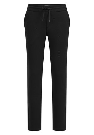 Kind geht und trägt eine schwarze Anzugjacke, ein gemustertes Hemd mit Fliege, schwarze Hose und weiße Turnschuhe vor einem einfachen hellen Hintergrund.