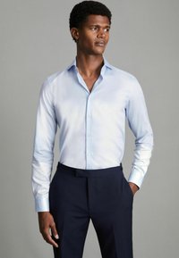 Chemise bleu clair à boutons en tissu lisse et léger, dotée d'un col classique, de manches longues et d'une coupe ajustée, associée à un pantalon sombre.