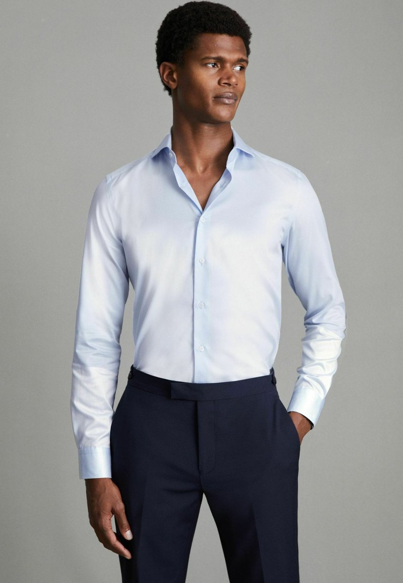 Chemise bleu clair à boutons en tissu lisse et léger, dotée d'un col classique, de manches longues et d'une coupe ajustée, associée à un pantalon sombre.