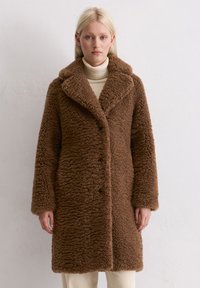 Cappotto marrone fluffy con una superficie texturizzata, colletto ampio e bottoni lungo la parte anteriore. Indossato sopra un dolcevita crema. Maniche lunghe.