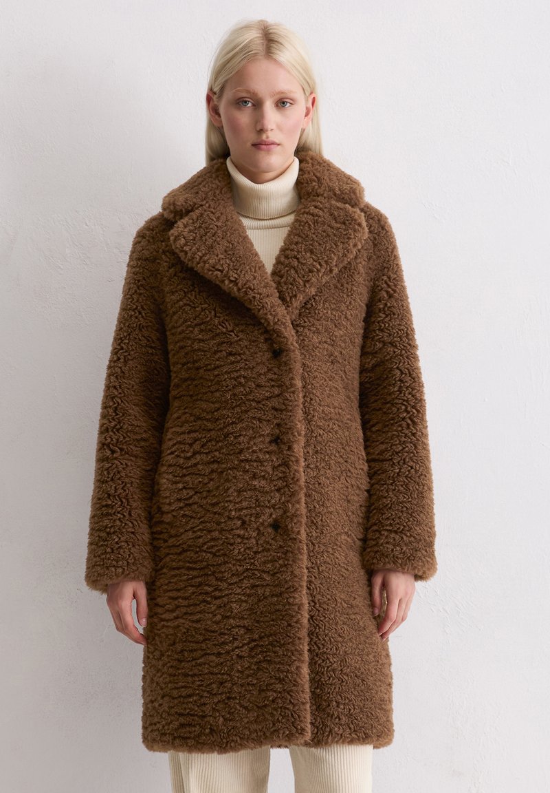 Cappotto marrone fluffy con una superficie texturizzata, colletto ampio e bottoni lungo la parte anteriore. Indossato sopra un dolcevita crema. Maniche lunghe.