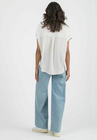 White short-sleeved blouse with back pleat, paired with light blue wide-leg trousers and white sneakers on a neutral background.