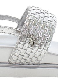 Stříbrné pletené sandály s širokým páskem, které mají hladkou texturu a jemný lesk. Podešev je bílá s mírným špalíčkem.