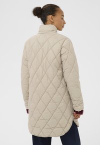 Beige quilted lang frakke med høj krave, der præsenterer diamantmønstersting og en let buet kant. Blød tekstur og afslappet design.