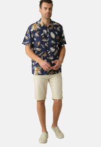 Korte mouwen marineblauw shirt met een palmbladpatroon in wit en oranje. Gecombineerd met beige shorts en lichte instap schoenen.