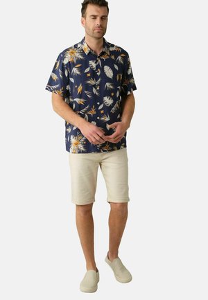 Chemise à manches courtes bleu marine avec un motif de feuilles de palmier en blanc et orange. Associée à un short beige et des chaussures de type slip-on légères.