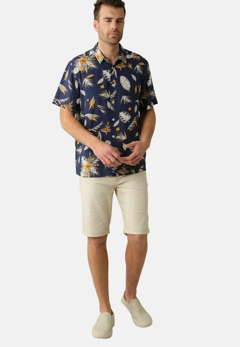Korte mouwen marineblauw shirt met een palmbladpatroon in wit en oranje. Gecombineerd met beige shorts en lichte instap schoenen.
