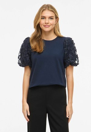 Femme portant un chemisier bleu marine à manches bouffantes texturées et un pantalon noir, debout devant un fond clair uni.