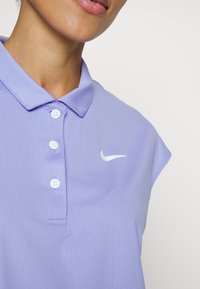 Gros plan d'une personne portant un polo Nike violet clair avec trois boutons et un logo swoosh blanc sur le côté droit de la poitrine.
