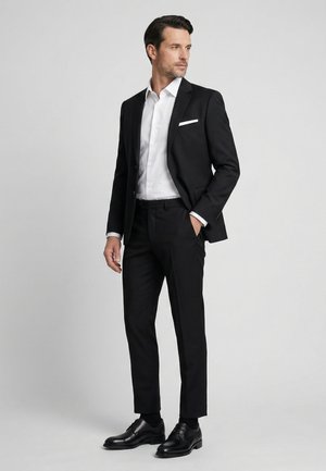 Mand i sort jakkesæt med hvid skjorte, sorte sko og hvid lommetørklæde, der ser til siden mod en ensfarvet baggrund.