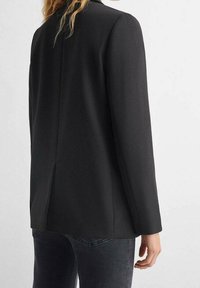 Black tailored blazer with long sleeves, a single back vent, and smooth texture, paired with dark denim trousers.