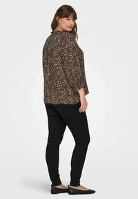 Moteris, vilkinti juodas liesas kelnes, juodus batus ir ilgą rudos leopardų rašto palaidinę, žiūri per petį prieš baltą foną.