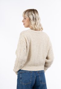 Beige gyapjú pulóver, rövid fazonnal, bordás alján és virágmintás hímzéssel az ujjakon; kék farmernadrággal párosítva.