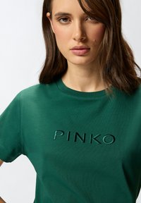 Zeleni pamukasti T-shirt s okruglim izrezom i izvezenim logotipom "PINKO" u istoj boji, s kratkim rukavima i glatkom teksturom.