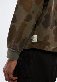 Χέρι ατόμου δίπλα σε φούτερ με καμουφλάζ μοτίβο, φθαρμένη μανσέτα και μικρή λευκή ετικέτα, σε συνδυασμό με μαύρο παντελόνι που έχει πλαϊνή τσέπη με κουμπί.