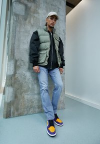 Olivo žalia ir juoda pūkinė liemenė uždėta ant juodos striukės, šviesiai mėlyni džinsai ir purpurinės/oranžinės sportbačiai su juodais raišteliais. Tekstūruotas betono fonas.