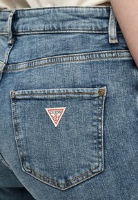 Πίσω τσέπη μπλε τζιν με κεντημένη στο κέντρο κόκκινη και λευκή τριγωνική ετικέτα λογότυπου Guess.