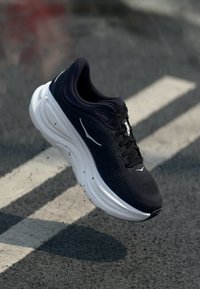 Chaussure de sport noire avec une tige en mesh texturé, une semelle intermédiaire blanche et un renfort au talon. Comprend des lacets et un logo sur le côté.