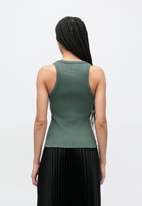 Canottiera verde a coste con design aderente, caratterizzata da spalline larghe e schiena a scollo tondo. Indossata sopra una gonna nera plissettata.