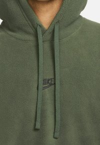 Olivgrüner Fleece-Hoodie mit einer Fronttasche, verstellbaren Kordeln und einem schwarzen gestickten Logo auf der Brust. Weicher Textur.
