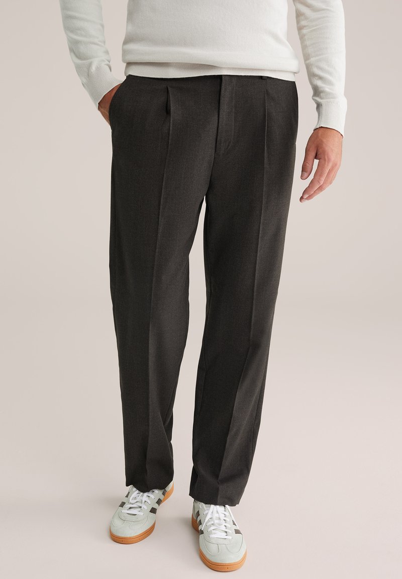 Sötétszürke redőzött nadrág, amely anyagból készült; fehér sportcipővel párosítva, szürke csíkokkal és barna talppal; világos pulóverrel viselve.