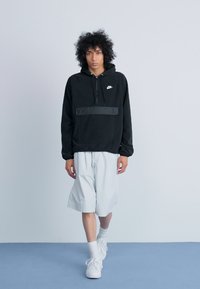 Črna kapuca Nike s sprednjim žepom, v kompletu s svetlo sivimi kratkimi hlačami. Model nosi bele superge in nogavice do gležnja. Enostavna ozadje.