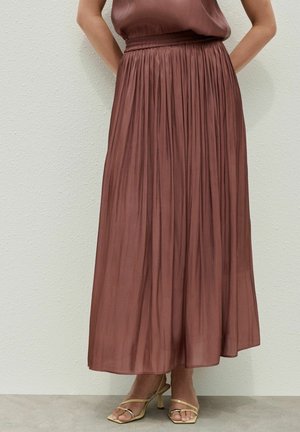Femme portant une jupe midi plissée marron taille haute associée à des sandales beige à talons ouverts, contre un mur uni.