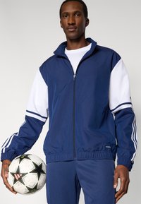 Giacca sportiva blu navy con accenti e strisce bianche, zip frontale e polsini elasticizzati. Il modello tiene una palla da calcio bianca e nera con motivo a fantasia.