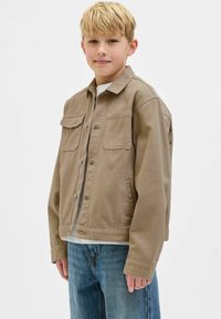 Khaki farmerdzseki, plüss gallérral, gombos elülső részel, két mellzsebbel és cuffolt hosszú ujjakkal, kék farmernadrággal párosítva.