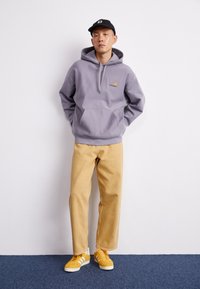 Šedá mikina s kapucí a přední kapsou, béžové manšestrové kalhoty a žluté tenisky s bílými pruhy, nošené s černou čepicí.