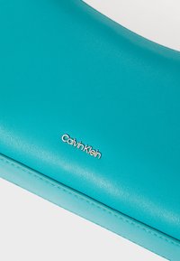 Turkos läderclutch i en slät textur med silverfärgad "Calvin Klein"-logotyp. Tillverkad av mjukt material, med en elegant design.