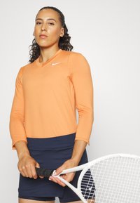 Top deportivo de manga larga naranja con escote en V, combinado con una falda azul marino. La modelo sostiene una raqueta de tenis blanca.