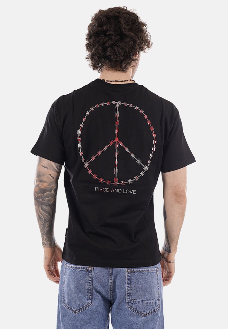 Uomo con capelli ricci che indossa una maglietta nera con un simbolo di pace fatto di filo spinato rosso e bianco e la scritta "PEACE AND LOVE" sul retro, jeans blu sbiaditi.