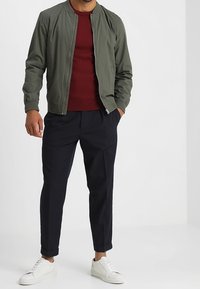 Olivgrön bomberjacka med dragkedja fram, bärs över en bordeauxröd tröja, kombinerad med marinblå avsmalnande byxor och vita sneakers.