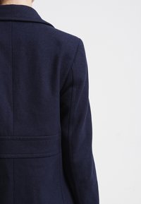 Manteau en laine marine avec un design structuré, doté d'un col cranté et d'une fente au dos. Texture lisse et silhouette ajustée.