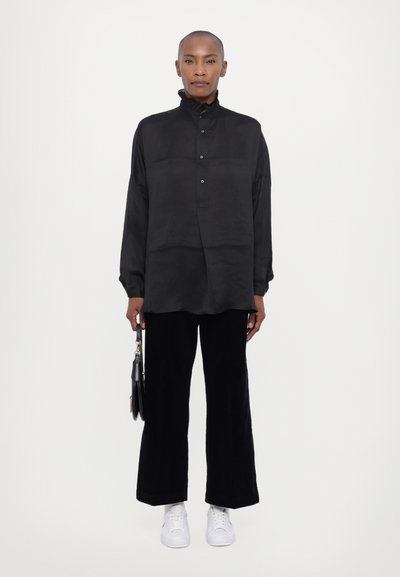 Fekete gombos ing rátétes gallérral, fekete kordbársony nadrággal és fehér tornacipővel párosítva. A modell egy kis fekete táskát tart a kezében.