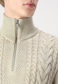 Maglione beige lavorato a maglia con colletto alto, dotato di una zip anteriore e motivi a treccia lungo le spalle. Tassello morbido.