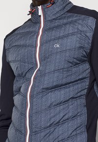 Navy and gray puffer jacket with herringbone pattern, zip front, high collar, and contrasting navy sleeves. White zipper with red accent.