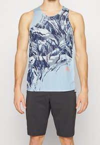 Top deportivo de color azul claro con un patrón abstracto en navy y blanco, con una etiqueta en la parte inferior derecha y un cuello redondo. Combinado con pantalones cortos negros.